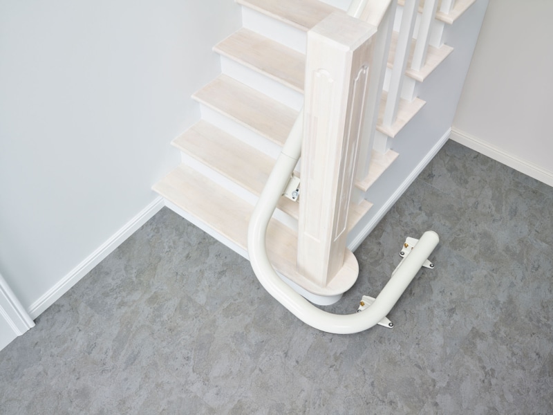 schienenlauf-am-gelaender-mit-bodenende-um-ecke Kurvige weiße Treppenliftschiene auf weißer Treppe I TK Home Solutions