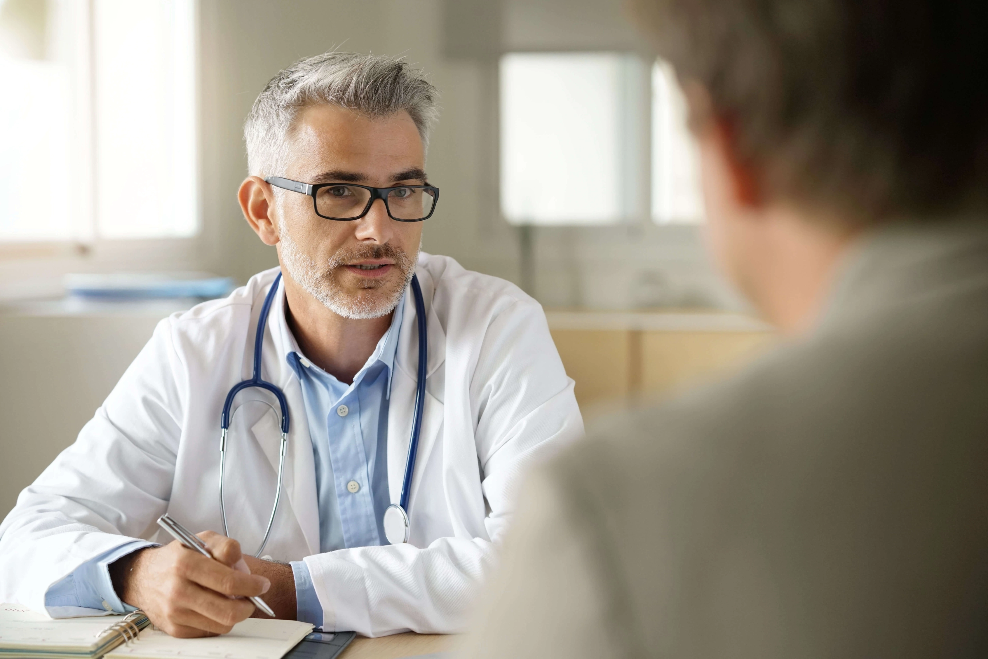 Ein Arzt schreibt in ein Notizbuch während eines Gespräches mit einem Patienten. Der Arzt ist im Fokus der Kamera, die hinter dem Patienten platziert ist | TK Home Solutions