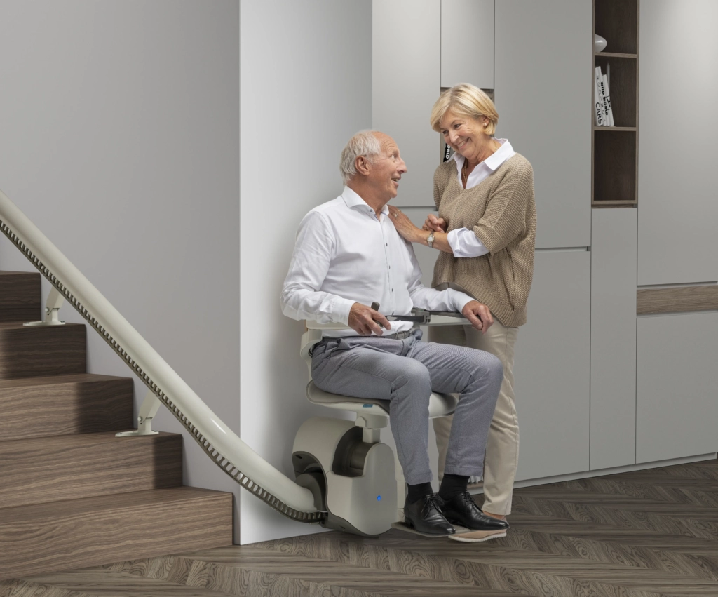 mann-sitzend-auf-treppenlift-frau-daneben-stehend Ein Mann sitzt auf dem Treppenlift und schaut sich dabei gegenseitig mit seiner danebenstehende Frau an | TK Home Solutions