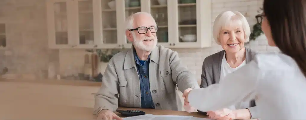 Header_Bild_seniorenpaar-bei-einer-vertragsschli Ein Seniorenpaar bei einer Vertragschließung mit einem Handschlag am Küchentisch. Die Kamera ist von vorne auf das Seniorenpaar gerichtet | TK Home Solutions