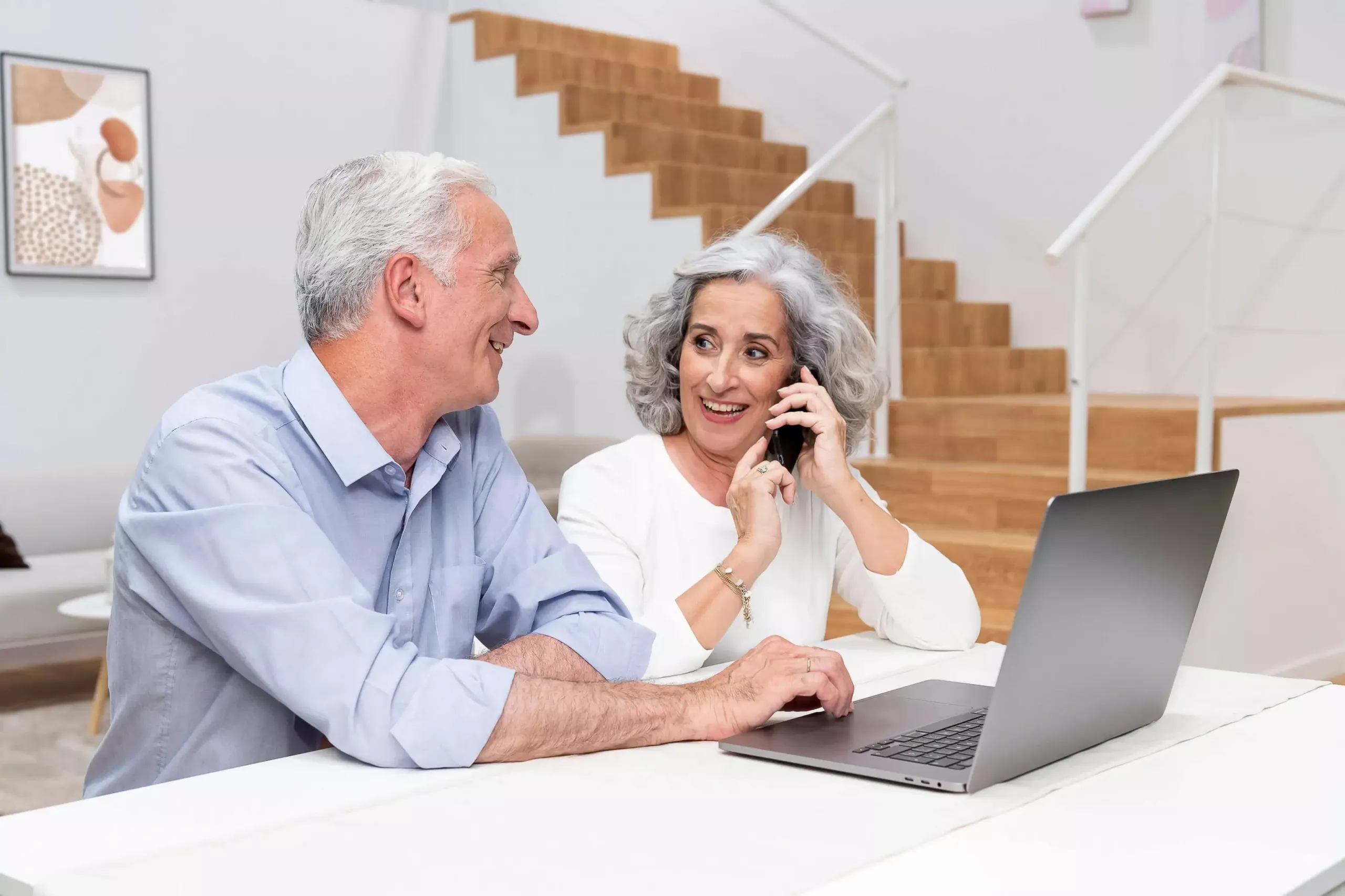 Ein älteres Paar informiert sich online an ihrem Laptop über Treppenlifte. Das Paar sieht sich dabei an und die Frau telefoniert währenddessen | TK Home Solutions