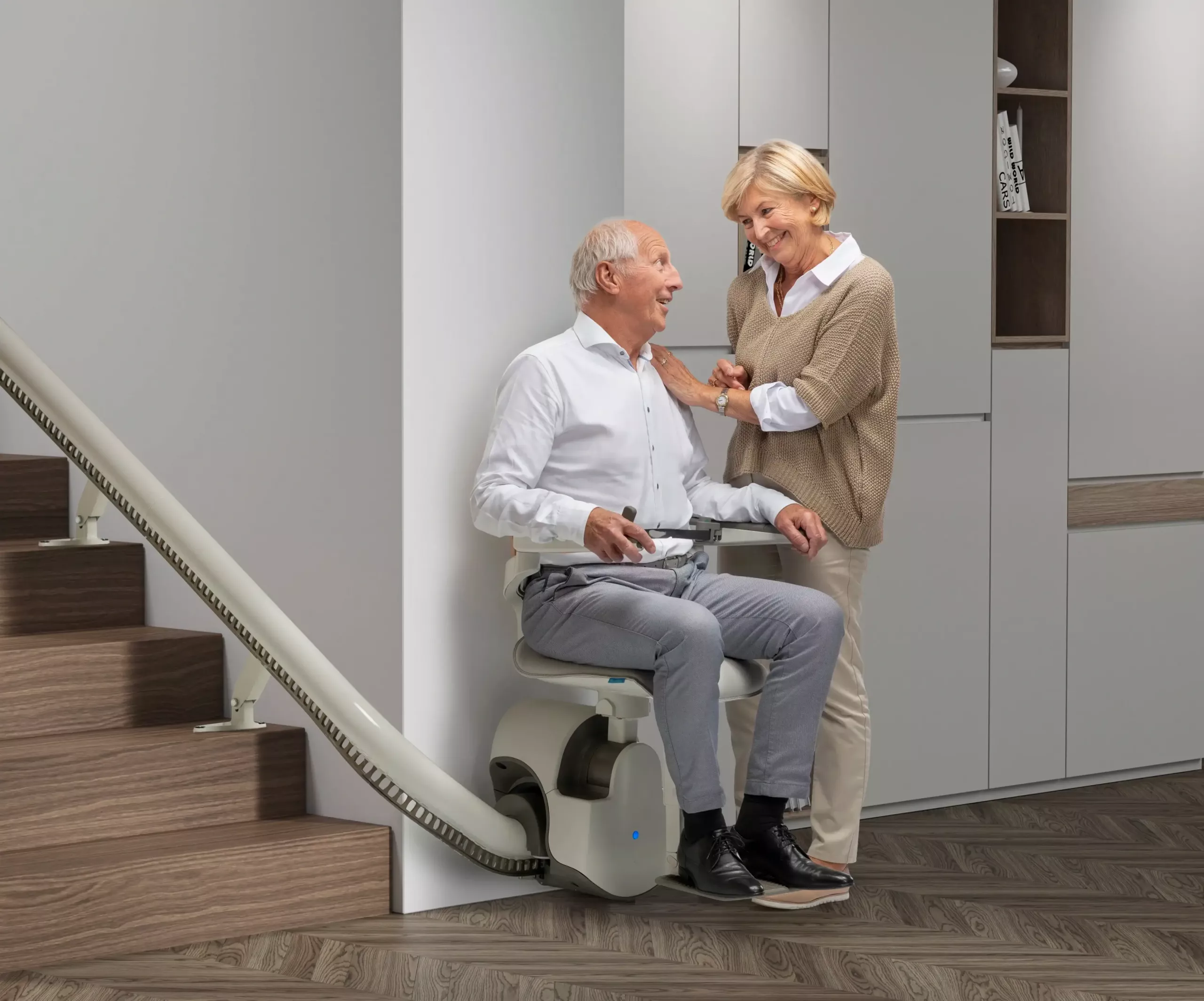 Ein Mann sitzt auf dem Treppenlift und schaut sich dabei gegenseitig mit seiner danebenstehende Frau an | TK Home Solutions