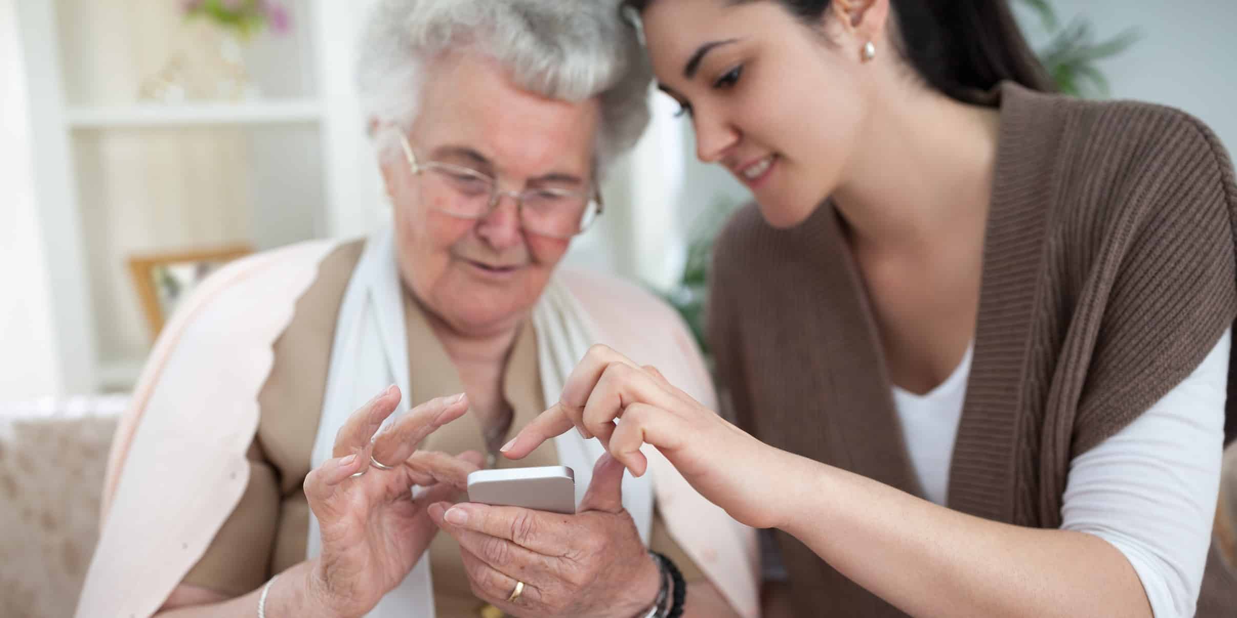AdobeStock_111227709 Cropped Eine junge Frau hilft einer alten Frau bei er Bedienung ihres Smartphones