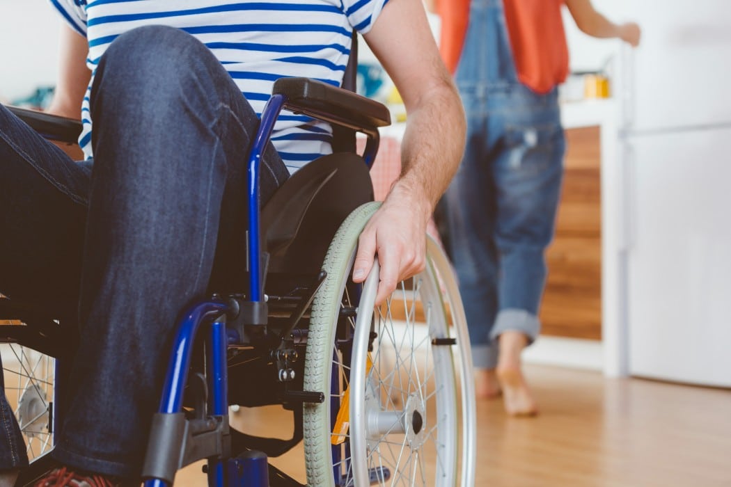 Person im Rollstuhl greift an das Rad; im Hintergrund steht eine weitere Person in einer Wohnung.