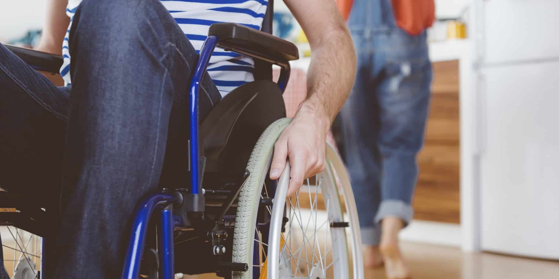 Header Lift Rollstuhlfahrer Person im Rollstuhl greift an das Rad; im Hintergrund steht eine weitere Person in einer Wohnung.